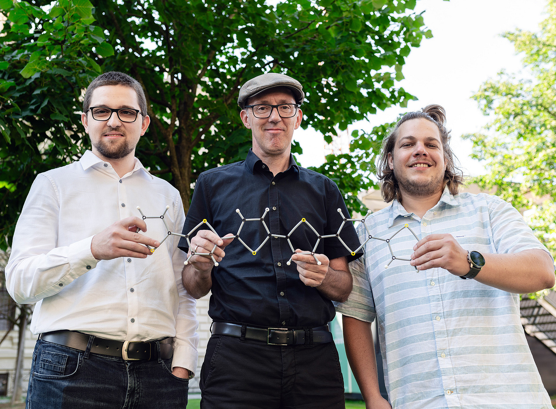 Three men of different ages, two wearing glasses, two in light-coloured shirts and one in a dark shirt, are standing outdoors in front of some trees, holding a chemical structure model between them. ©Uni Graz/Angele