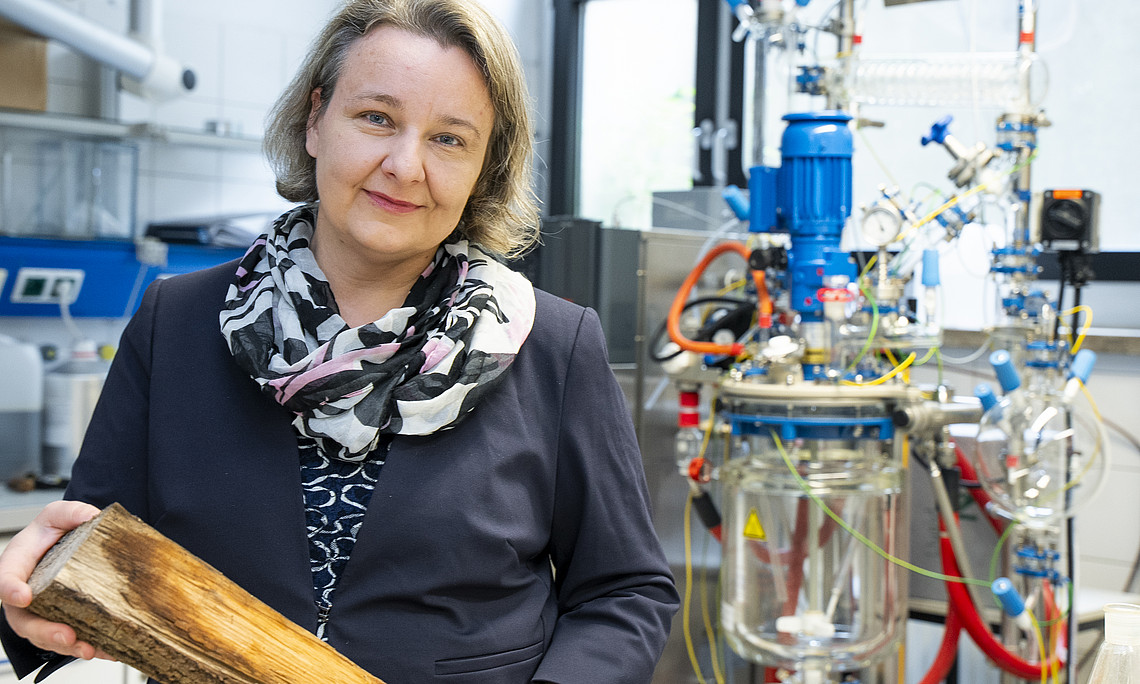 photograph of the scientist holds wood in her hand and stands next to an equipment with pipes, cables and glass bubble chamber at lab background, middle-aged woman wearing navy blue suit jacket with black patterned scarf around neck, short hair
