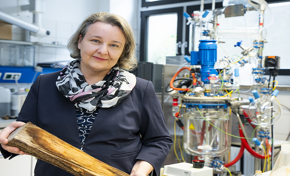 Foto einer Wissenschaftlerin, die Holz in der Hand hält und neben einem Gerät mit Rohren, Kabeln und einer Glasblasenkammer im Labor steht, mittlere Frau in marineblauer Anzugjacke mit schwarz gemustertem Schal um den Hals, kurze Haare