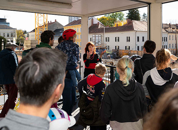 Architektin Hemma Fasch steht mit Schüler:innen auf der Terrasse des Info-Points. ©Uni Graz/Tzivanopoulos