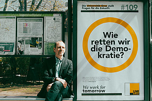 Florian Bieber an einer Bushaltestelle vor einem Plakat mit seiner Forschungsfrage Wie retten wir die Demokratie?