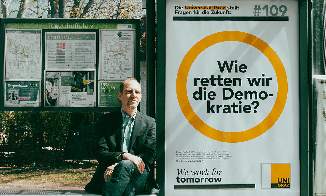 Florian Bieber an einer Bushaltestelle vor einem Plakat mit seiner Forschungsfrage Wie retten wir die Demokratie?