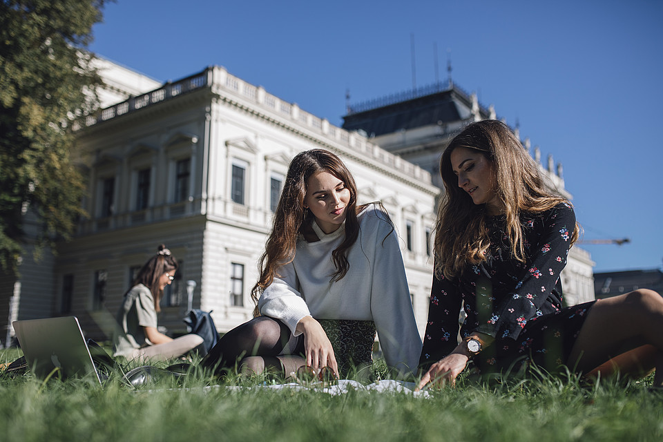 Zwei Studierende unterhalten sich sitzend auf der Campuswiese vor dem Hauptgebäude