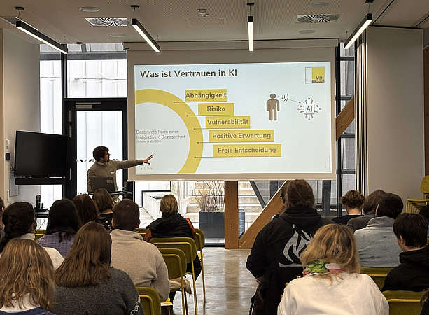 ein Mann steht vor einem Tisch mit Laptop und hält einen Vortrag, hinter ihm sieht man die Leinwand mit der Präsentation. Im Publikum sitzen viele Menschen und hören zu ©Uni Graz/Schmieder