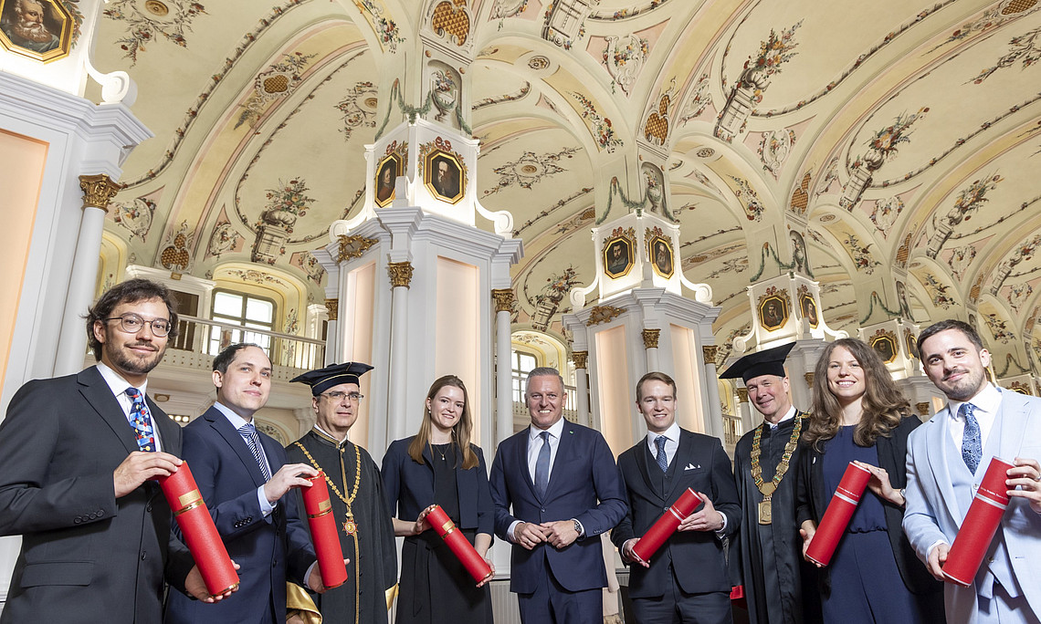 Eine Gruppe festlich gekleideter Personen steht in einer prunkvollen, hellen Halle mit hoher, reich bemalter Decke und großen weißen Säulen mit goldenen Verzierungen. Mehrere Personen tragen dunkle Talare und akademische Kopfbedeckungen, die übrigen Anzüge oder elegante Kleider. Alle halten rote, zylindrische Urkundenrollen in den Händen. Im Hintergrund sind hohe Fenster, Galerien und weitere architektonische Details des Saales zu erkennen.