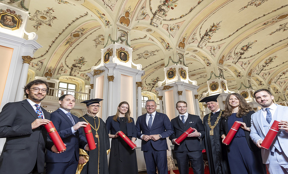 Eine Gruppe festlich gekleideter Personen steht in einer prunkvollen, hellen Halle mit hoher, reich bemalter Decke und großen weißen Säulen mit goldenen Verzierungen. Mehrere Personen tragen dunkle Talare und akademische Kopfbedeckungen, die übrigen Anzüge oder elegante Kleider. Alle halten rote, zylindrische Urkundenrollen in den Händen. Im Hintergrund sind hohe Fenster, Galerien und weitere architektonische Details des Saales zu erkennen.