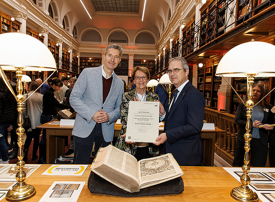 Zwei Männer und eine Frau halten eine Urkunde in der Hand. Vor Ihnen auf einem Tisch ein altes Buch ©Uni Graz/Thomas Luef