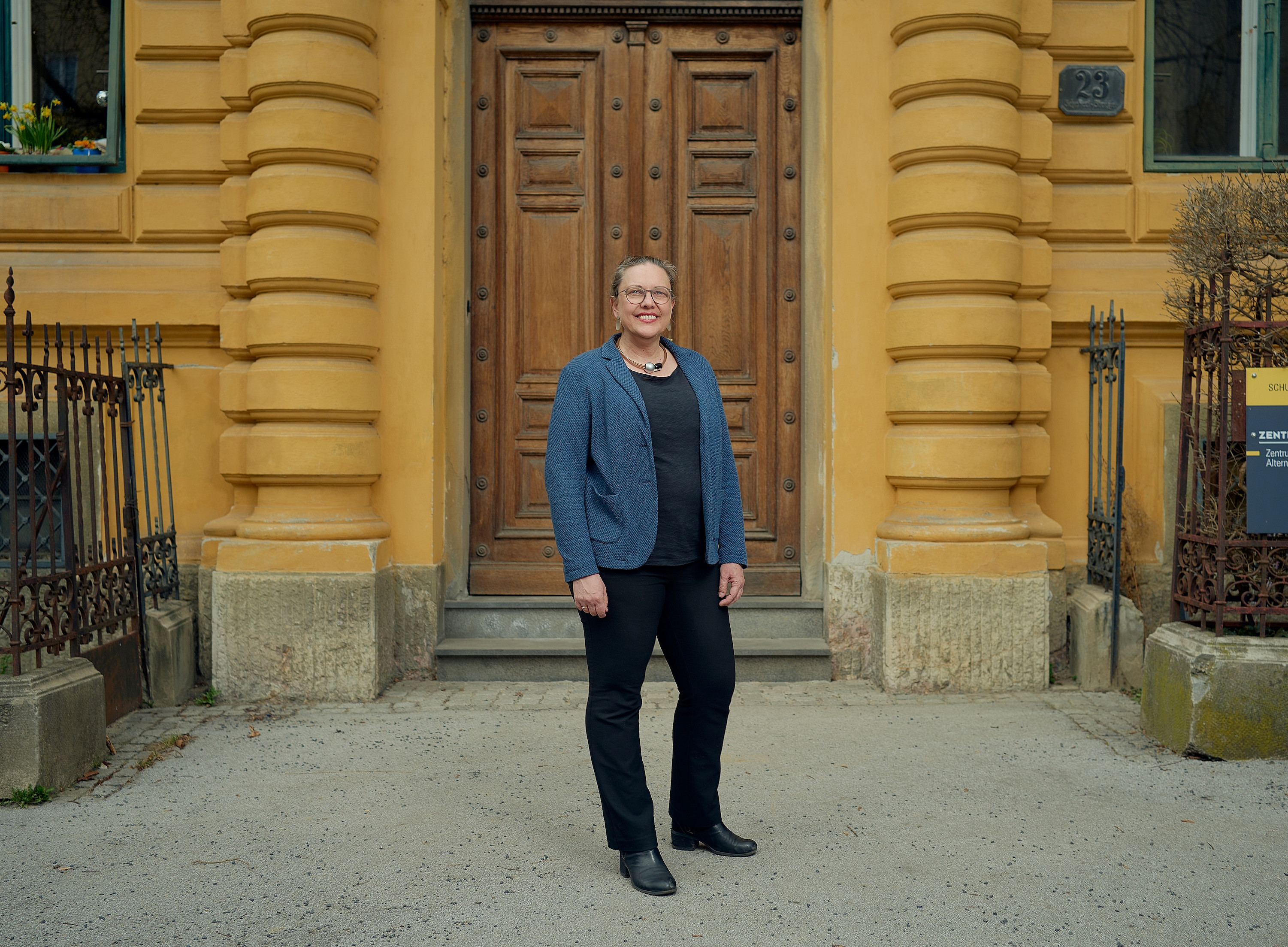 Eine Frau steht vor einem Gebäude mit einer beigen Fassade. Sie hat einen blauen Blazer an 