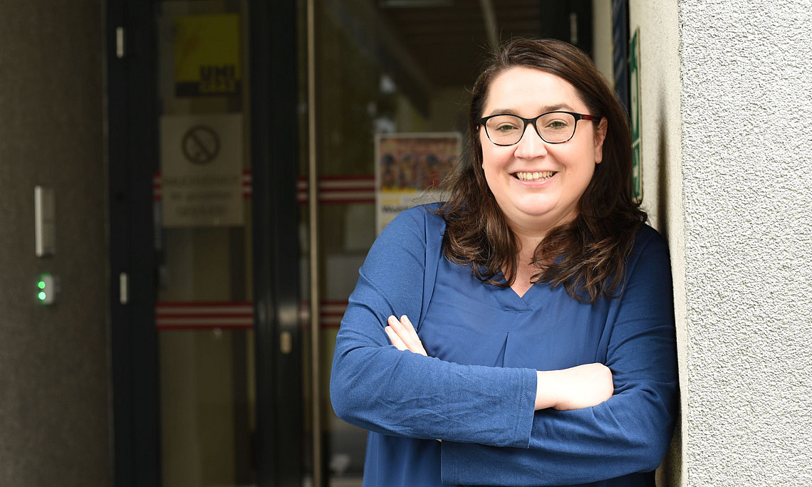 A photograph of a woman with shoulder-length brown hair and glasses, wearing a blue top, standing in front of the entrance to her office and smiling at the camera. She is leaning against the wall outside the door. 