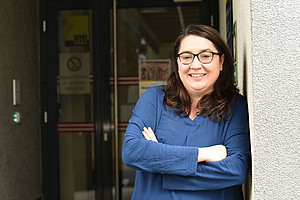 Ein Foto einer Frau mit schulterlangen braunen Haaren und Brille, die ein blaues Oberteil trägt und vor dem Eingang zu ihrem Büro steht und in die Kamera lächelt. Sie lehnt an der Wand vor der Tür. 