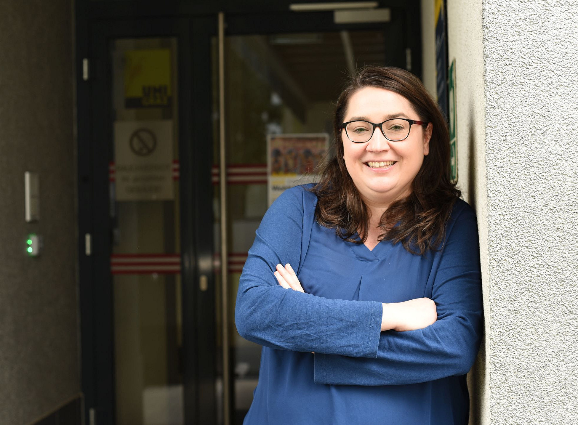 Ein Foto einer Frau mit schulterlangen braunen Haaren und Brille, die ein blaues Oberteil trägt und vor dem Eingang zu ihrem Büro steht und in die Kamera lächelt. Sie lehnt an der Wand vor der Tür. 