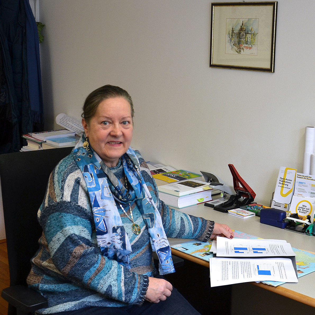 eine Frau mit Pullover und Halstuch sitzt an einem Schreibtisch vor Dokumenten