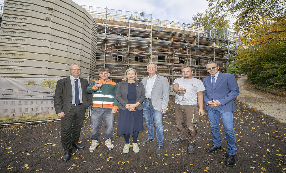 Ein Gruppenfoto von Regierungsbeamten mit sechs Personen, die lächelnd vor einem im Bau befindlichen unfertigen Haus stehen. Zwei Arbeiter in Wohnbauuniformen halten ein Glas Wein in der Hand. Der Hintergrund ist mit beigen Betonplatten und grauen Metallfliesen verkleidet. Im Hintergrund sind grüne Bäume hinter der Straße zu sehen.