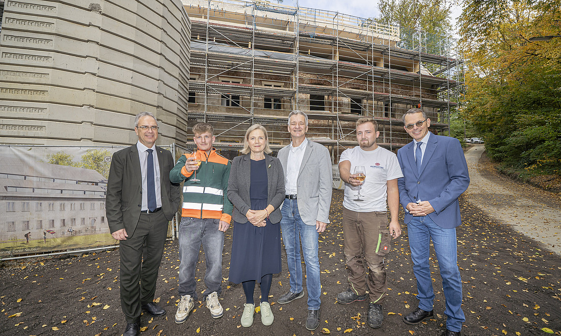 Ein Gruppenfoto von Regierungsbeamten mit sechs Personen, die lächelnd vor einem im Bau befindlichen unfertigen Haus stehen. Zwei Arbeiter in Wohnbauuniformen halten ein Glas Wein in der Hand. Der Hintergrund ist mit beigen Betonplatten und grauen Metallfliesen verkleidet. Im Hintergrund sind grüne Bäume hinter der Straße zu sehen.