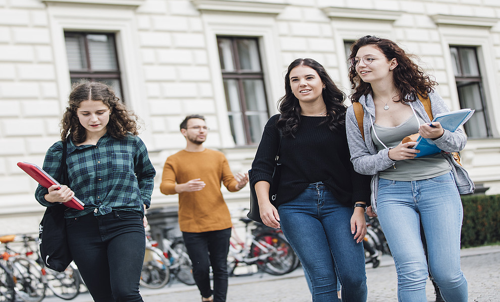 Drei junge Frauen mit Mappen in den Händen stehen vor einem alten Gebäude. Im Hintergrund ist auch ein Mann zu erkennen und Fahrradständer