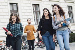 Three young women with folders in their hands are standing in front of an old building. In the background, a man and a bicycle stand can also be seen.