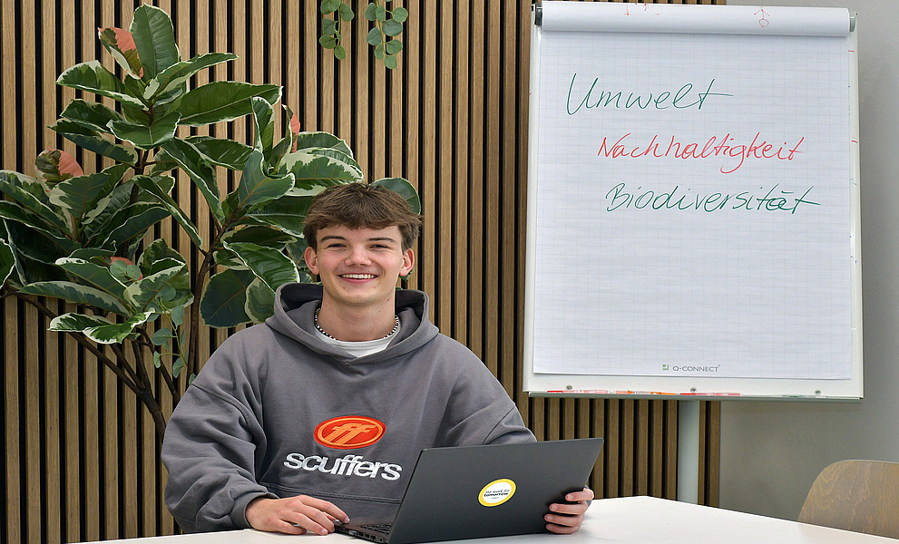 Valentin Happe mit einem Laptop an einem Tisch sitzend, im Hintergrund eine Pflanze und ein Flipchart, darauf die Worte "Umwelt", "Nachhaltigkeit", "Biodiversität"