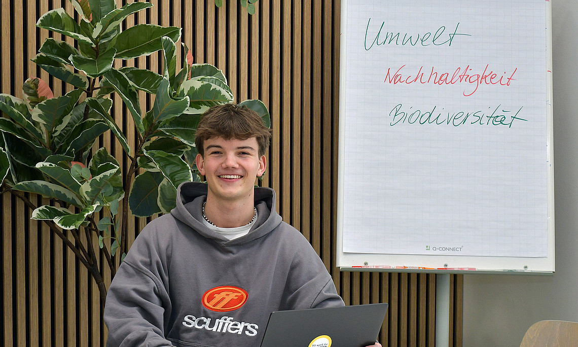 Valentin Happe sitting at a table with a laptop, in the background a plant and a flipchart with the words "Umwelt" (environment), "Nachhaltigkeit" (sustainability), "Biodiversität"(biodiversity) 