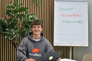 Valentin Happe sitting at a table with a laptop, in the background a plant and a flipchart with the words "Umwelt" (environment), "Nachhaltigkeit" (sustainability), "Biodiversität"(biodiversity) 