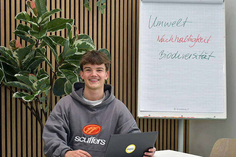 Valentin Happe sitting at a table with a laptop, in the background a plant and a flipchart with the words "Umwelt" (environment), "Nachhaltigkeit" (sustainability), "Biodiversität"(biodiversity) 