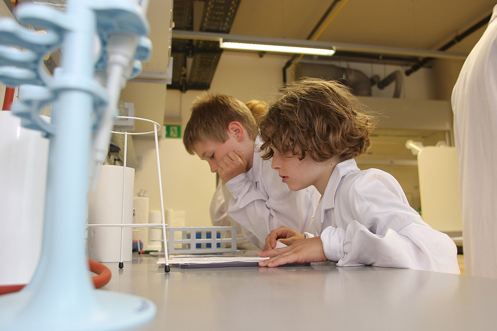 drei Kinder in weißen Labormänteln sind über einen Labortisch gebeugt und lesen eine Versuchsanleitung im Offenen Labor ©Grumet Lukas