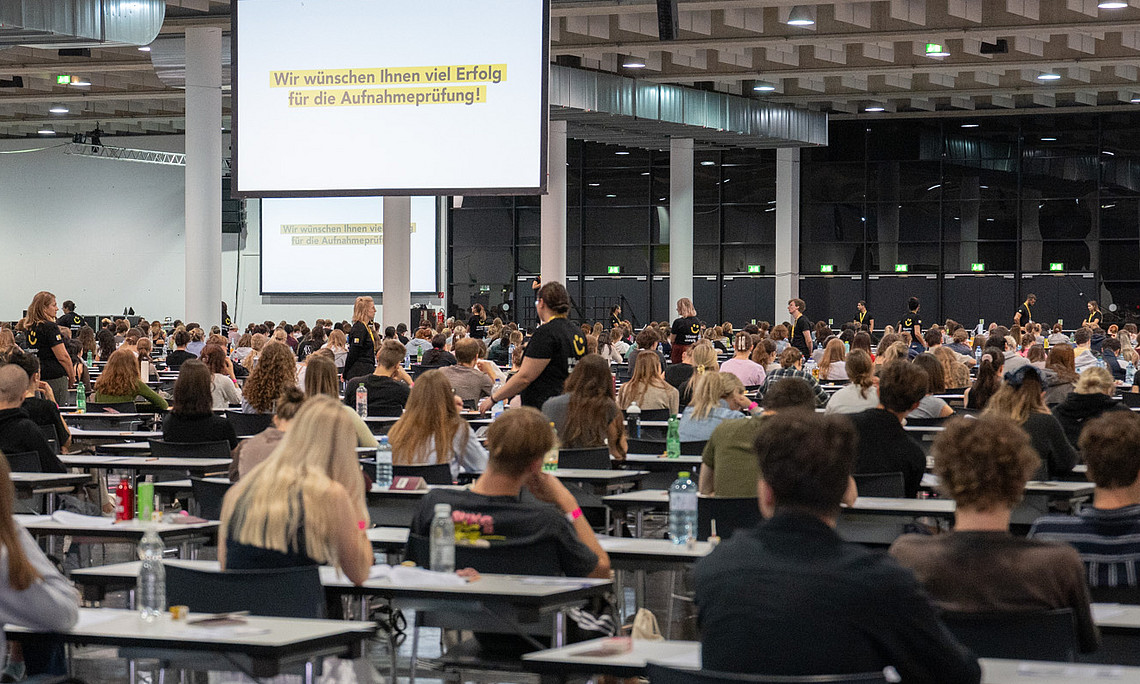 Große Prüfhalle mit dicht besetzten Einzeltischen, an denen zahlreiche Personen einen Aufnahmetest schreiben; hinten Leinwand mit eingeblendeter Begrüßungs- und Erfolgsbotschaft, einige Aufsichtspersonen bewegen sich zwischen den Reihen.