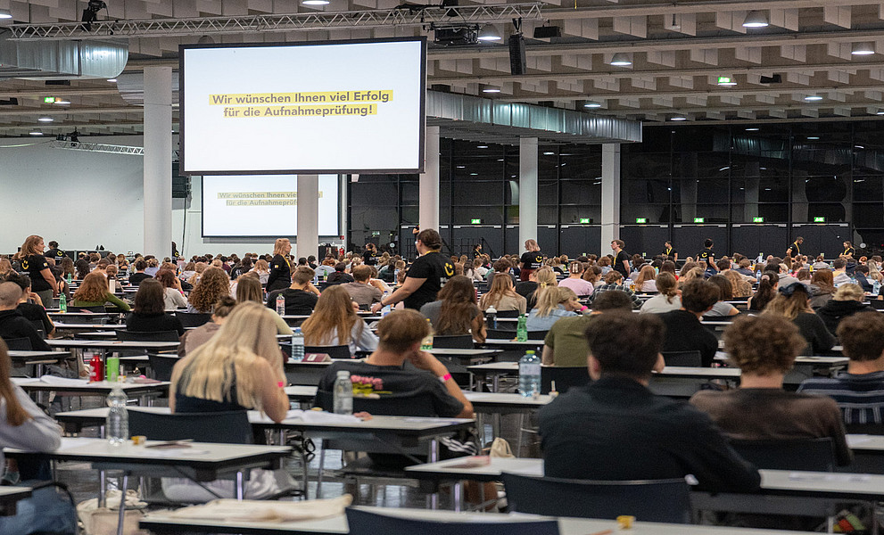 Große Prüfhalle mit dicht besetzten Einzeltischen, an denen zahlreiche Personen einen Aufnahmetest schreiben; hinten Leinwand mit eingeblendeter Begrüßungs- und Erfolgsbotschaft, einige Aufsichtspersonen bewegen sich zwischen den Reihen.