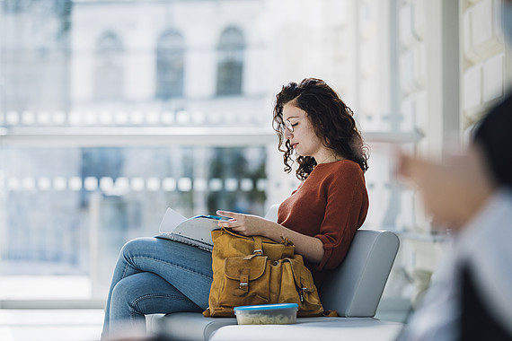 Studierende sitzt auf einer Bank im Innenbereich der Universität und liest in einem College-Block.