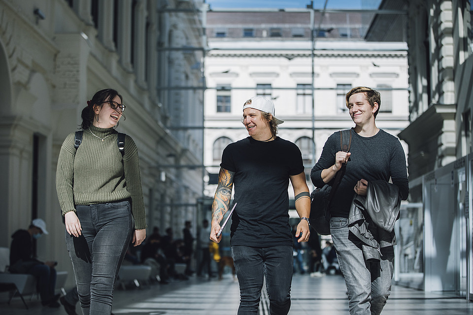 Studierende gehen lachend nebeneinander durch den offenen Indoor-Bereich der Universitätsbibliothek Graz.