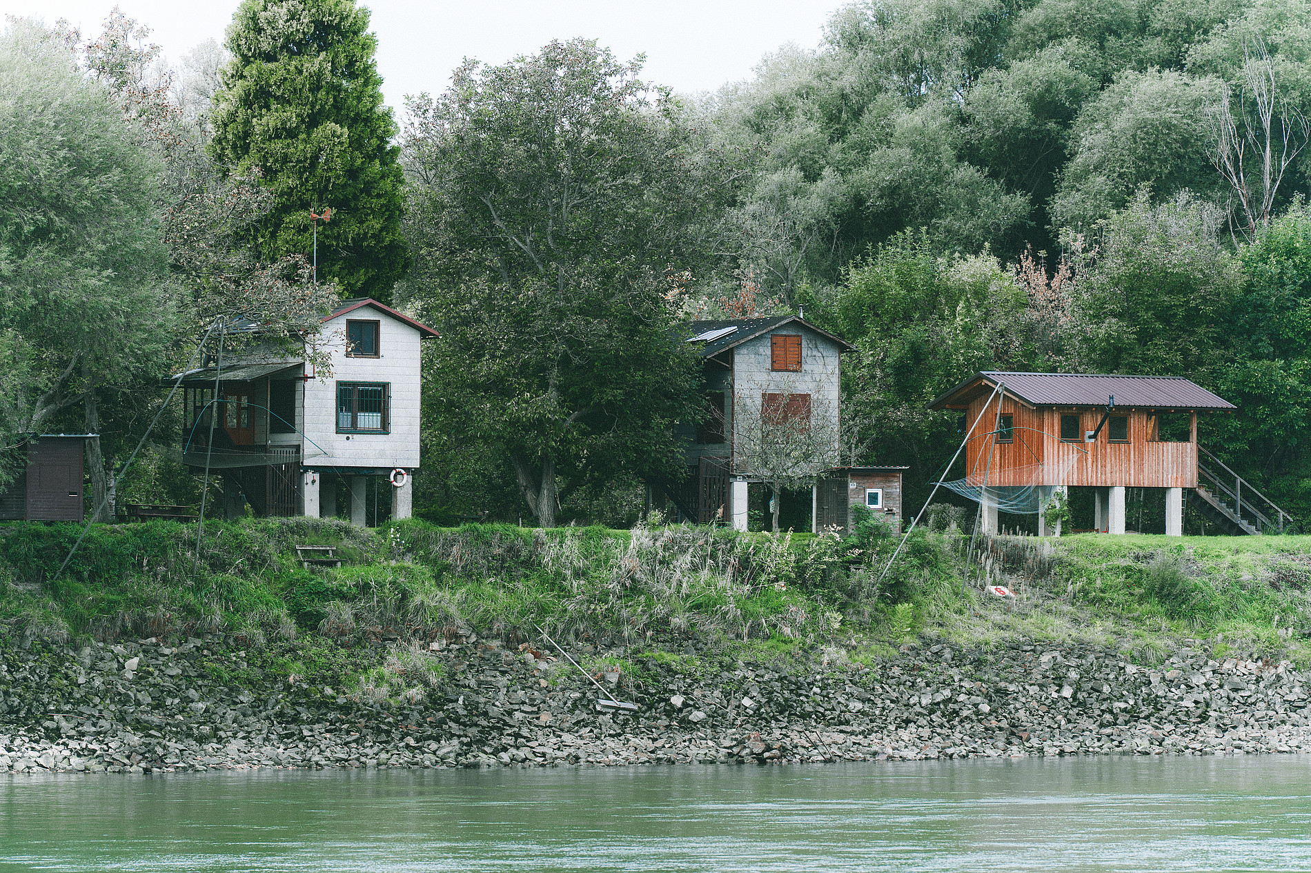 Three small houses on stilts along a riverbank, surrounded by dense green trees and vegetation. ©Manuel Madaini