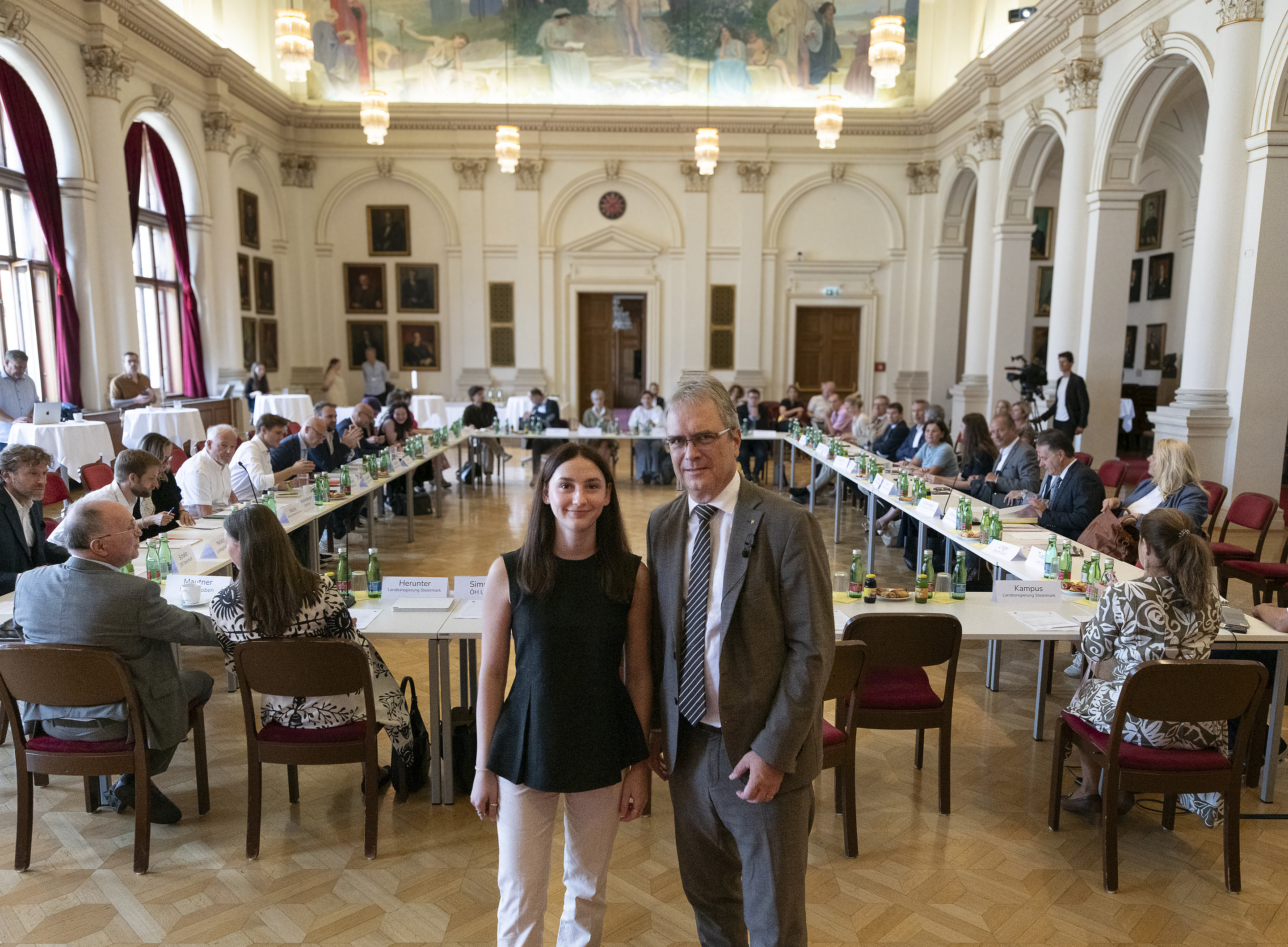 Ein Mann und eine Frau stehen in einem großen Saal, dahinter Menschen! ©Uni Graz/Tzivanopoulos