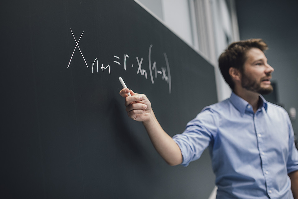 Professor steht vor der Tafel und zeigt mit der Kreide auf eine Formel, die er niedergeschrieben hat; er spricht dazu; sein Blick ist in den Hörsaal gerichtet
