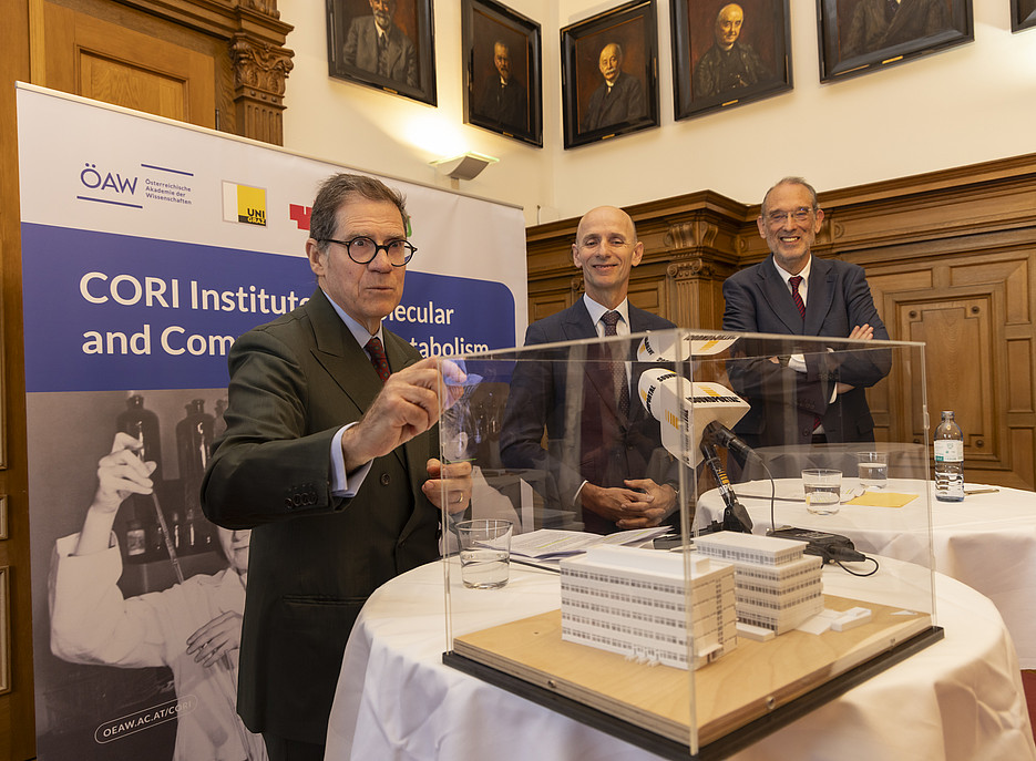 In a wood-panelled hall, three men of middle to advanced age are standing at two round, white-clothed bar tables. In the foreground on the left, a man in a dark suit is reaching for a transparent cover over a white model of a building, which is standing in a rectangular box on the table. Behind him, in the centre, stands a man in a suit and tie; to the right, another man in a suit with his arms crossed. Several microphones with white windscreens are positioned in front of the building model. In the background on the left is a large roll-up banner bearing the inscription ‘CORI Institute of Molecular and Computational Metabolism’. ©Helmut Lunghammer