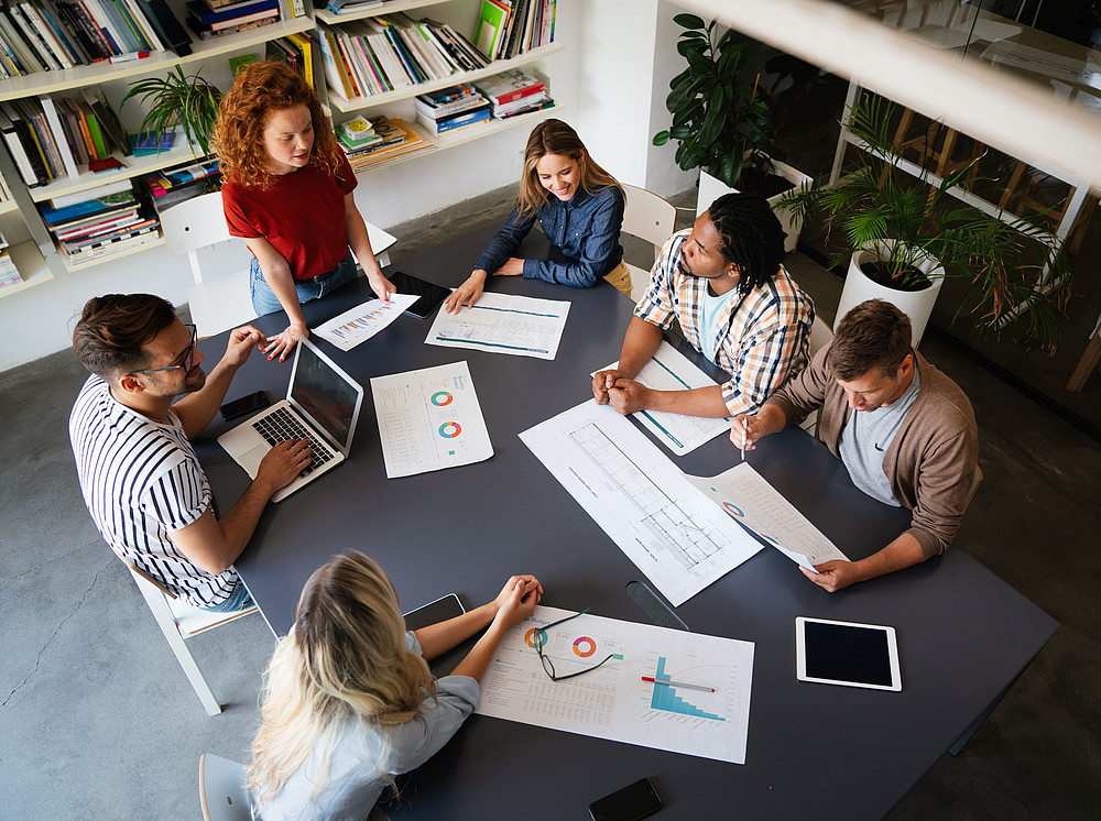 Gruppe von Personen arbeitet an einem Schreibtisch vor unterschiedlichen Unterlagen. ©NDABCREATIVITY - stock.adobe.com