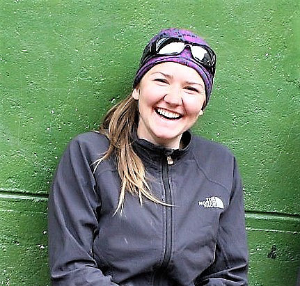 Fotografie von Geowissenschaften-Absolventin Katharina Brandner, wie sie lachend in Outdoor-Kleidung vor einer grünen Wand sitzt.