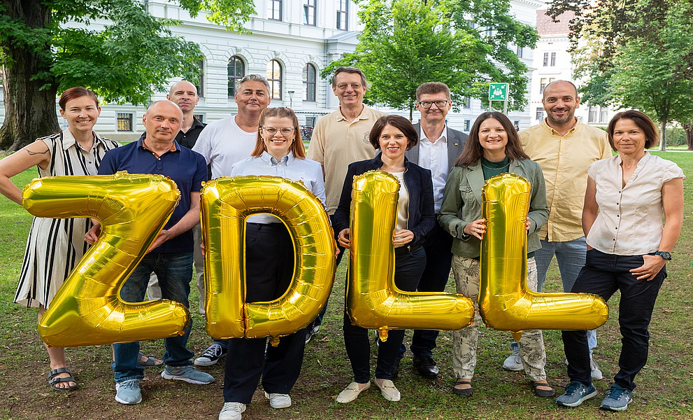 Eine Gruppe von Personen steht auf einer Wiese vor einem historischen Gebäude mit weißen Fassaden und großen Fenstern. Im Vordergrund halten einige von ihnen goldene Luftballons, die die Buchstaben 'ZDLL' formen. Im Hintergrund sind Bäume und ein gepflegter Park zu sehen.