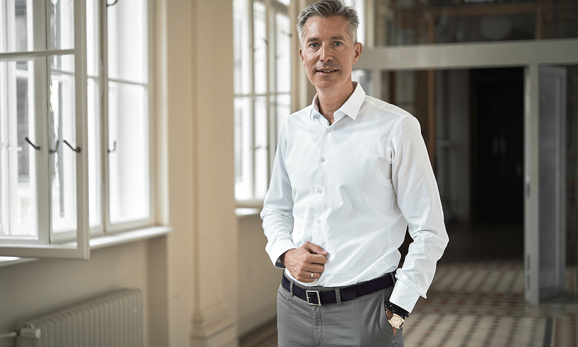 A person stands in a bright, elegant interior with large windows and a radiator on the wall. The floor is covered with patterned tiles, and doors and architectural details are visible in the background. The person is wearing a white shirt and gray pants.