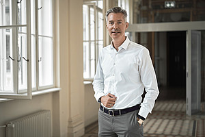 A person stands in a bright, elegant interior with large windows and a radiator on the wall. The floor is covered with patterned tiles, and doors and architectural details are visible in the background. The person is wearing a white shirt and gray pants.