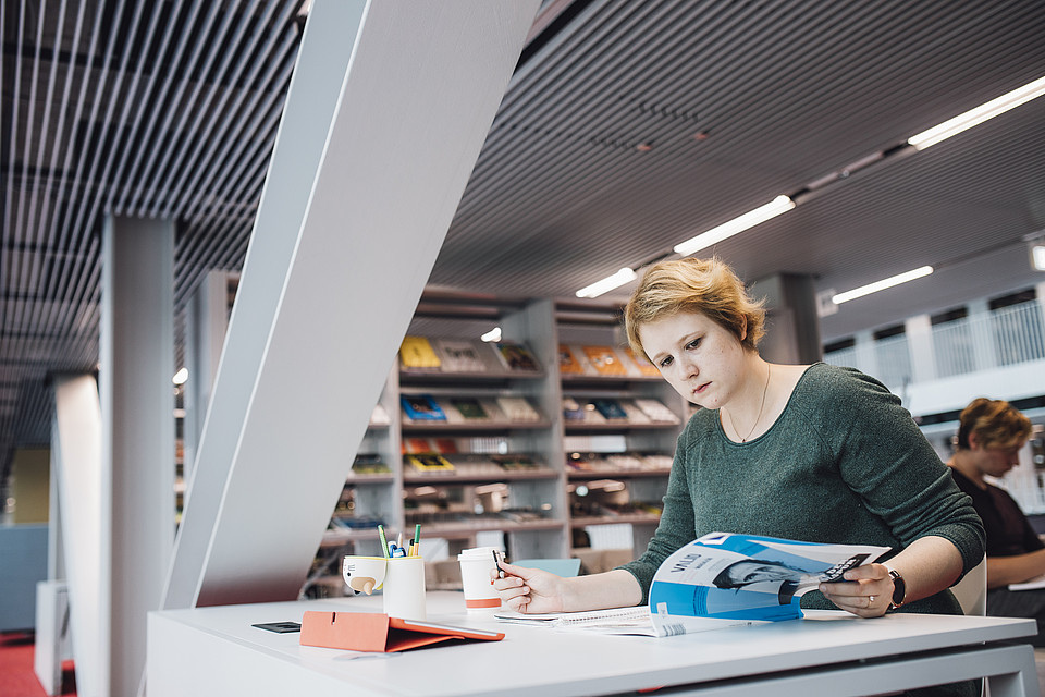 Eine Studierende lernt an einem Tisch in der Universitätsbibliothek und blättert in ihren Lernunterlagen
