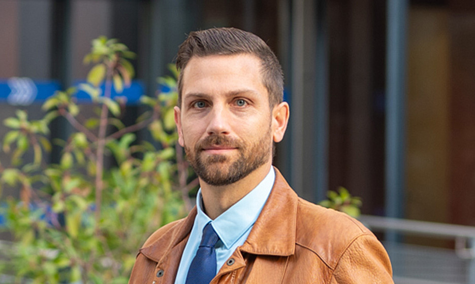 A man with short brown hair and a beard is standing outside a building. He is wearing a light blue shirt, a blue tie and a brown leather jacket. His hands are in his jacket pockets. In the background, you can see autumn leaves and a metal grille, as well as some plants and modern architecture. ©Uni Graz/Weber