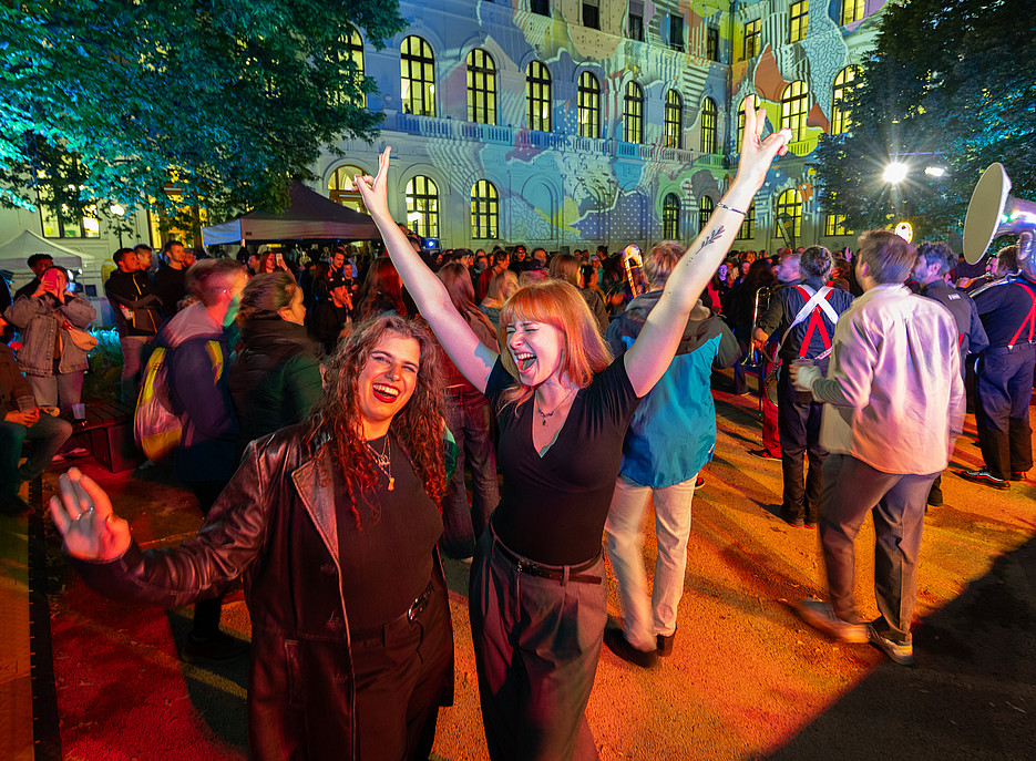 Menschen tanzen und feiern abends im Freien vor einem mehrstöckigen historischen Gebäude, das bunt mit Lichtmustern angestrahlt wird; im Vordergrund stehen zwei Personen mit erhobenen Armen, im Hintergrund sind weitere Feiernde und eine Blaskapelle mit glänzenden Instrumenten zu sehen, die Szene ist in kräftige Rot-, Blau- und Gelbtöne getaucht. ©Uni Graz/Harry Schiffer