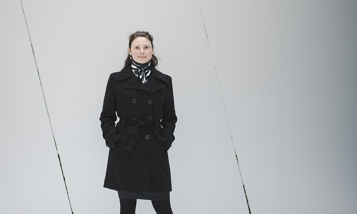 Person wearing a black coat, black tights and black shoes standing frontally on snow-covered ground in front of a large, light grey, slightly slanted segmented metal wall; hands in coat pockets, black scarf with white pattern, minimalist, cool background.