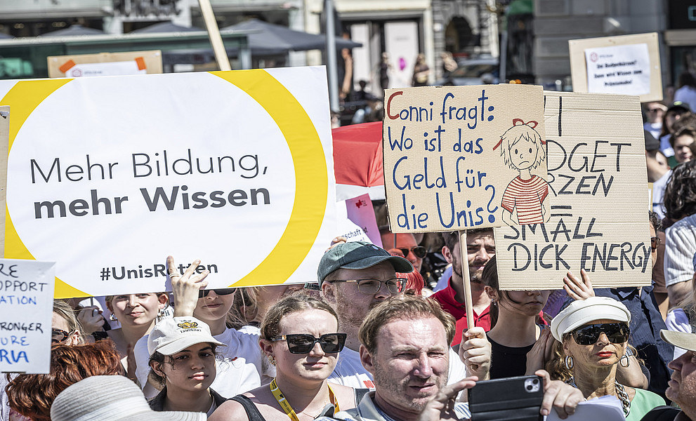 Menschenmenge bei einer Demo, viele junge Erwachsene halten Plakate mit Forderungen nach mehr Bildung und Geld für Universitäten hoch.