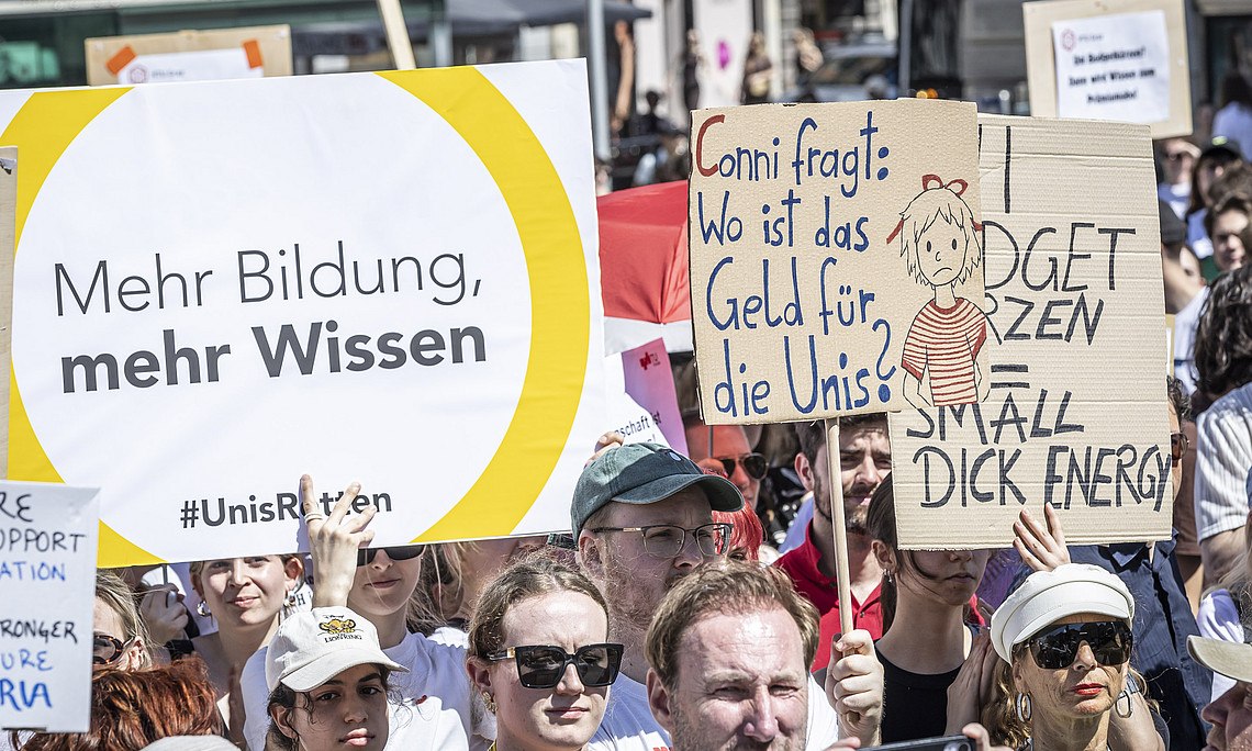 Menschenmenge bei einer Demo, viele junge Erwachsene halten Plakate mit Forderungen nach mehr Bildung und Geld für Universitäten hoch.