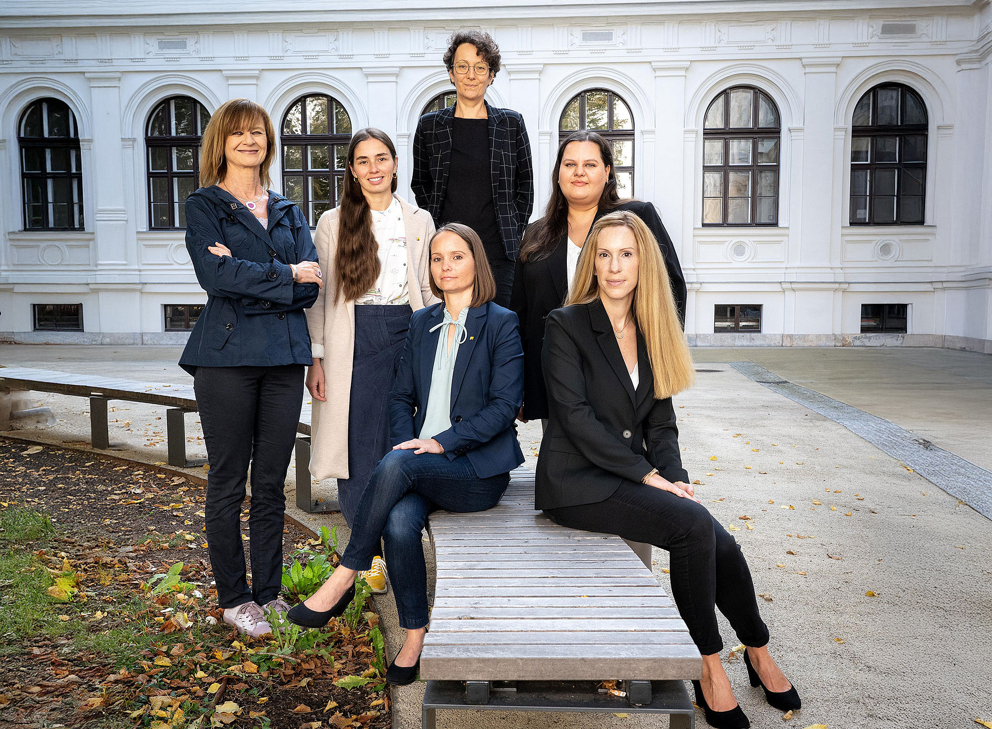 AKGL chairwoman Maria Elisabeth Aigner (left) and the AKGL Office team: Magdalena Halb, Ulrike Schustaczek, Carmen Klasicka (standing, from left), Johanna Kerschbaumer and Barbara Luttenberger (seated on a wooden bench, from left), in the courtyard of the main building of the University of Graz ©Alexandra Neubauer
