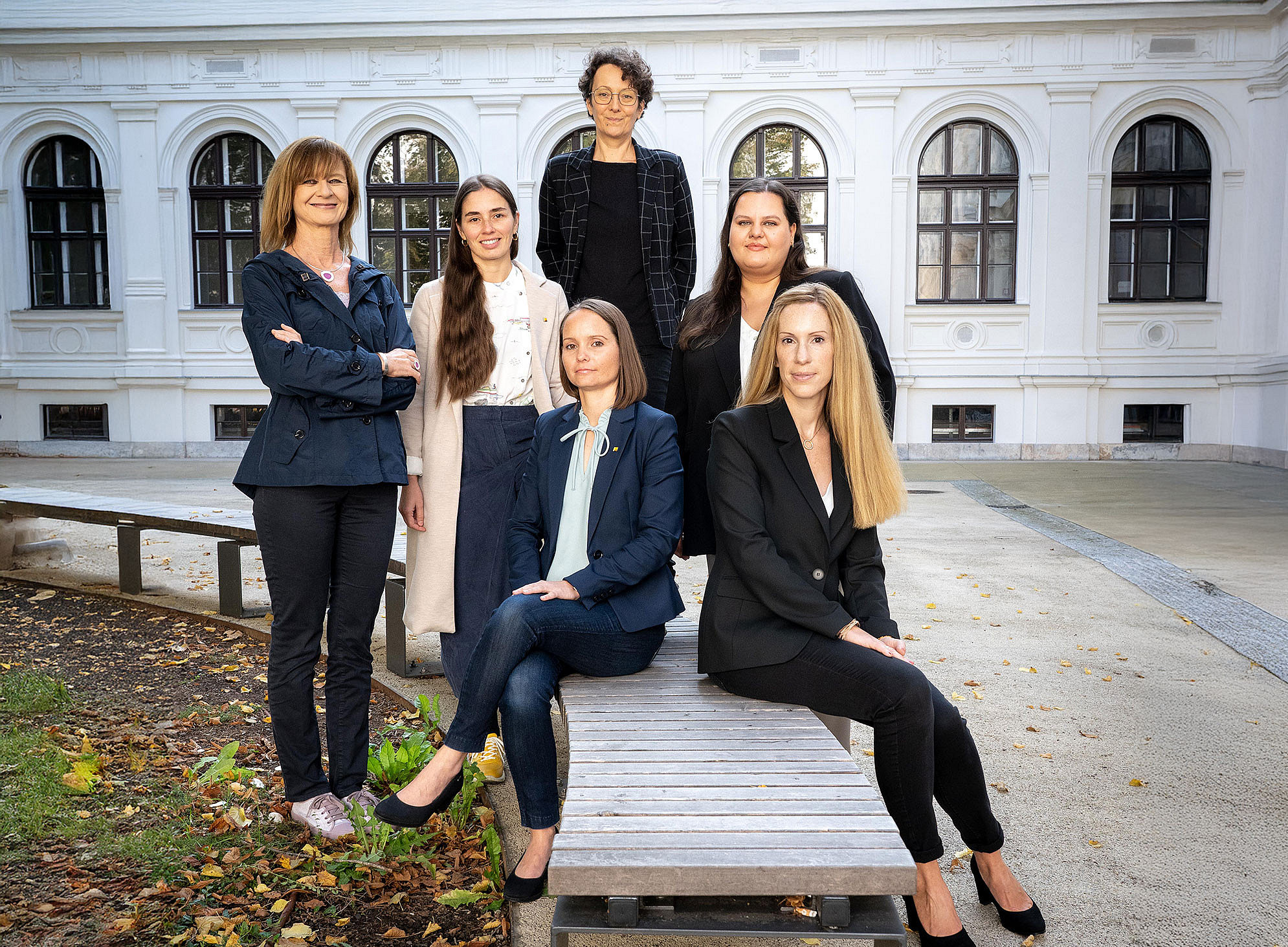 AKGL-Vorsitzende Maria Elisabeth Aigner (l.) mit dem Team des AKGL-Büros: Magdalena Halb, Ulrike Schustaczek, Carmen Klasicka (stehend v.l.), Johanna Kerschbaumer und Barbara Luttenberger (sitzend auf einer Holzbank v.l.), im Innenhof des Hauptgebäudes der Uni Graz ©Alexandra Neubauer