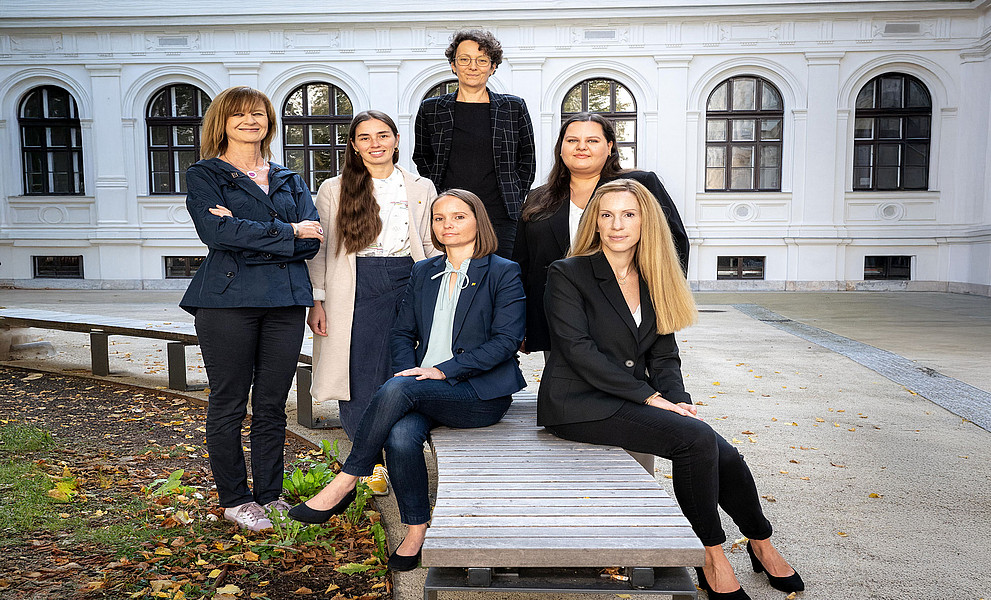 AKGL-Vorsitzende Maria Elisabeth Aigner (l.) mit dem Team des AKGL-Büros: Magdalena Halb, Ulrike Schustaczek, Carmen Klasicka (stehend v.l.), Johanna Kerschbaumer und Barbara Luttenberger (sitzend auf einer Holzbank v.l.), im Innenhof des Hauptgebäudes der Uni Graz