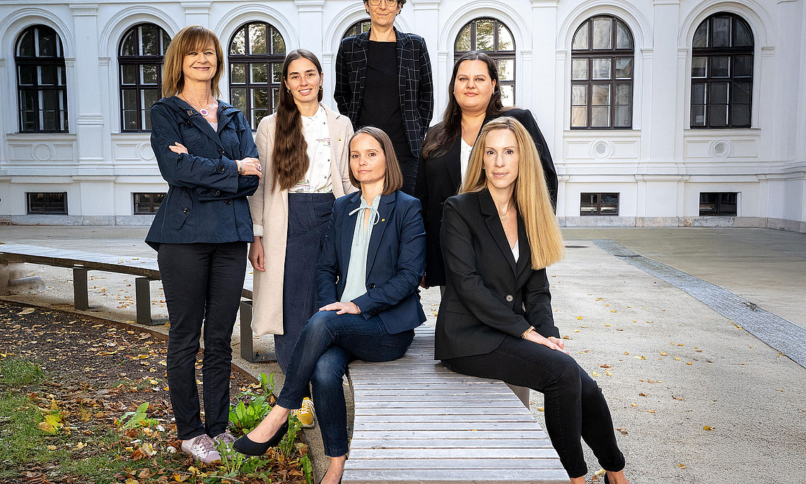 AKGL chairwoman Maria Elisabeth Aigner (left) and the AKGL Office team: Magdalena Halb, Ulrike Schustaczek, Carmen Klasicka (standing, from left), Johanna Kerschbaumer and Barbara Luttenberger (seated on a wooden bench, from left), in the courtyard of the main building of the University of Graz