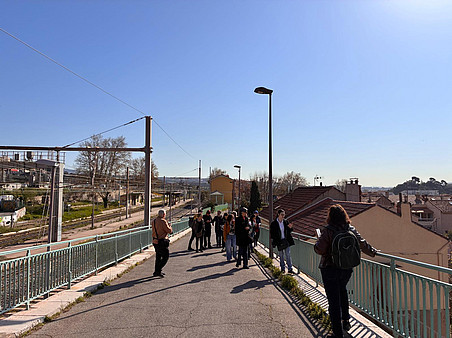 A group of people standing and walking along an elevated pedestrian path near a railway line, overlooking rooftops and tracks under a clear blue sky, with sunlight casting long shadows. ©Ben Gidley