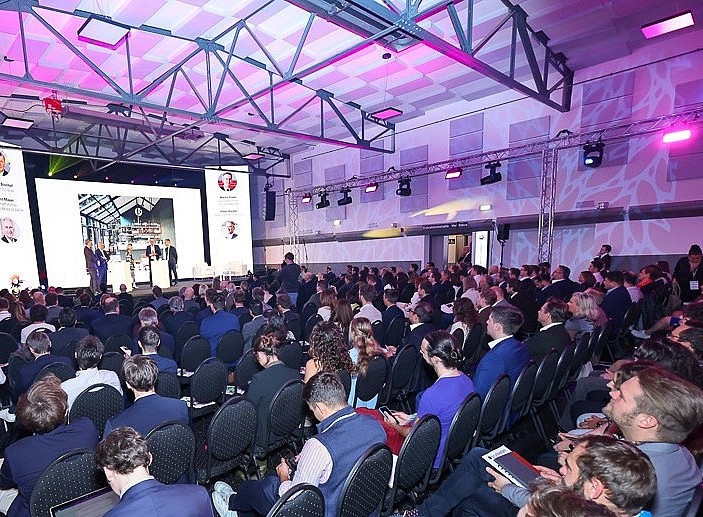 Eine Eventhalle voller Publikum, in der Ferne eine Bühne mit drei Personen und LED-Walls im Hintergrund ©Christian_Mikes